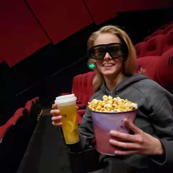 I am at the cinema wearing 3D glasses holding a bucket of popcorn and a fresh fruit drink 