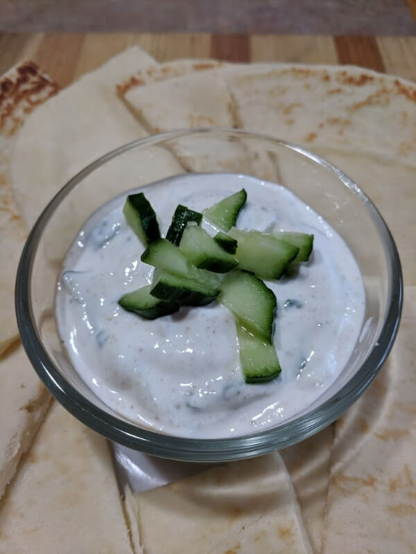 A bowl of cucumber raita surrounded by slices of naan bread.