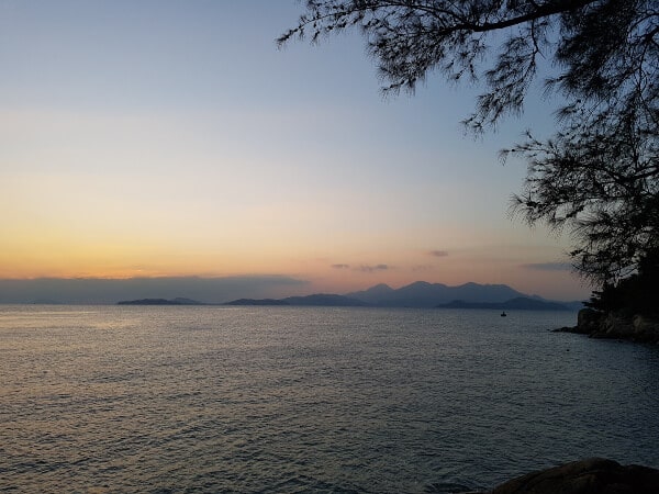 a view of the sky as the sun just set, there are mountains in the background and a tree frames the right side of the photo The sky is all different colors and the water is very still 