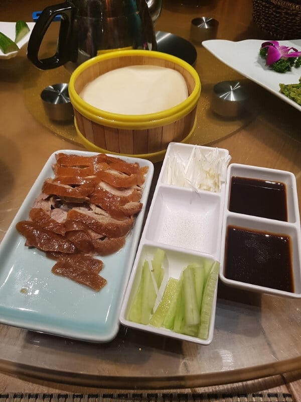 There is a tray of Peking duck, a dish of wraps and a tray with garnishes: sauces, cucumber and sugar