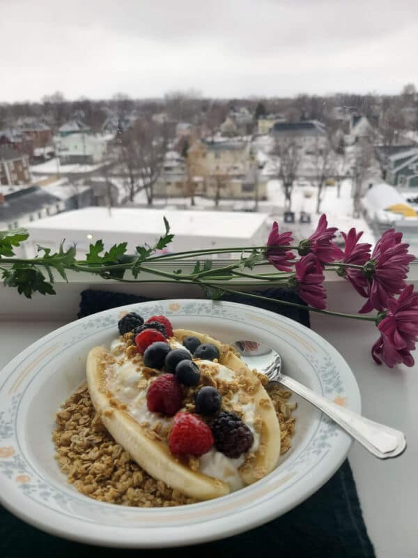 A yogurt banana split on a window ledge with a view of houses down below. Yogurt, a sliced banana, berries, and granola in a dish with a spoon and there are some pink flowers on the ledge beside the bowl. 