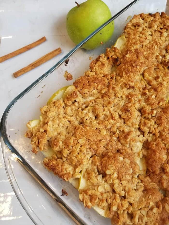 A pan of Low Sugar Apple Crisp Recipe with apples and cinnamon sticks beside it 