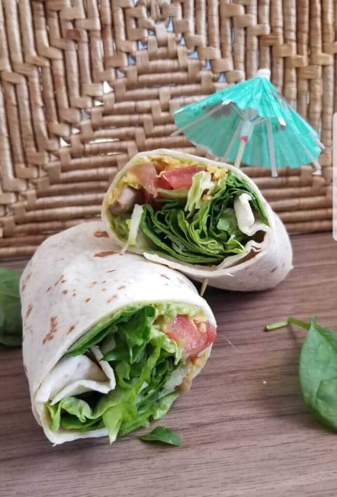 How to make Vegetarian Wraps Recipe: A wrap cut in half showing lettuce, spinach, cheese, tomatoes and other veggies. A drink umbrella is stuck in the top half of the wrap.