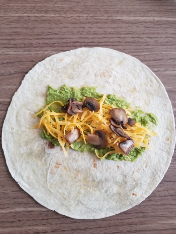 A wrap with avocado, lettuce, tomatoes, spinach, fried mushrooms and shredded cheese. 