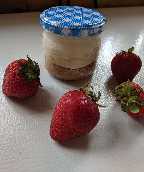 A jar filled with the graham crumbs and topped with cream cheese filling. There are some strawberries beside the jar.