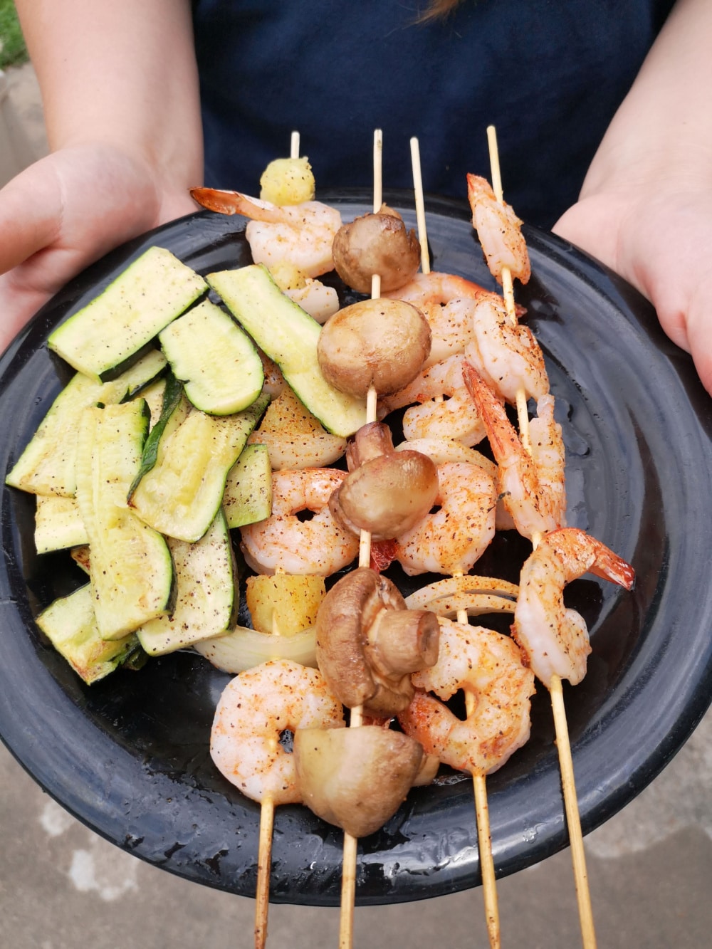 Someone is holding out a plate of grilled shrimp, mushrooms and zucchini. 