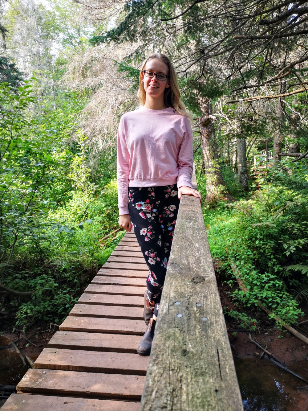 A girl is standing on a wooden bridge wearing flowered leggings and a pink sweater. There are trees and greenery all around me. 
