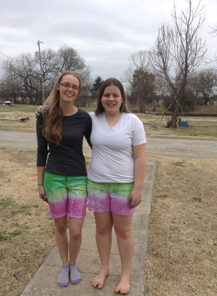 A photo of two girls wearing tie dyed shorts to look like watermelons. There is a strip of pink at the bottom with black dots to look like seeds, a thin strip of white and then different shades of green for the watermelon rind. 