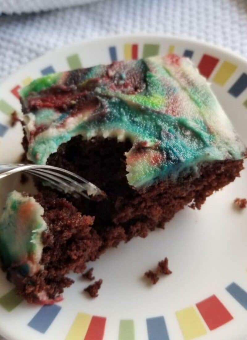 A slice of vegan chocolate cake decorated in colorful icing. There is a for taking a piece out of the slice of cake,