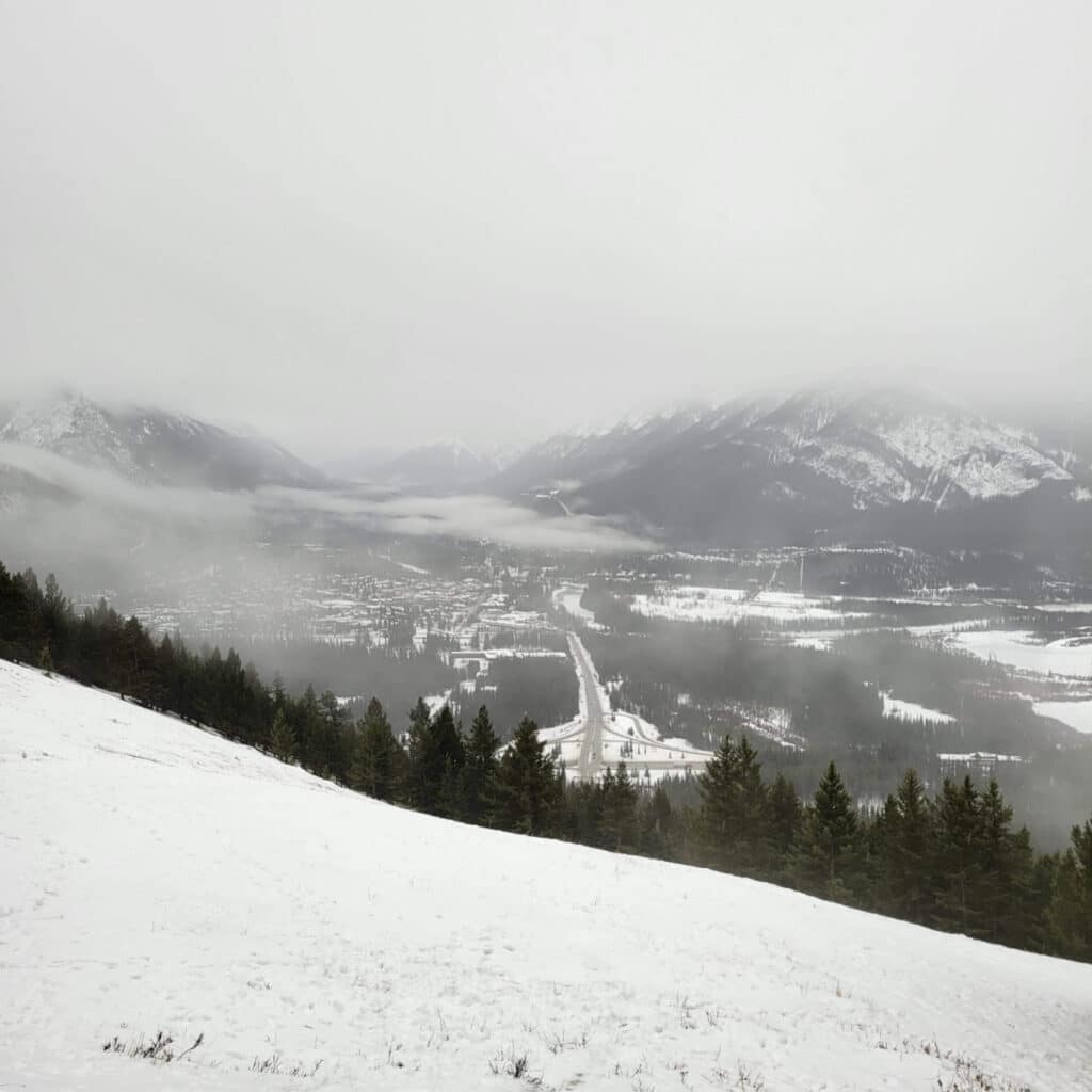 View from a lookout point. Below is: the town of Banff, mountains, trees, and there is mist above the town.