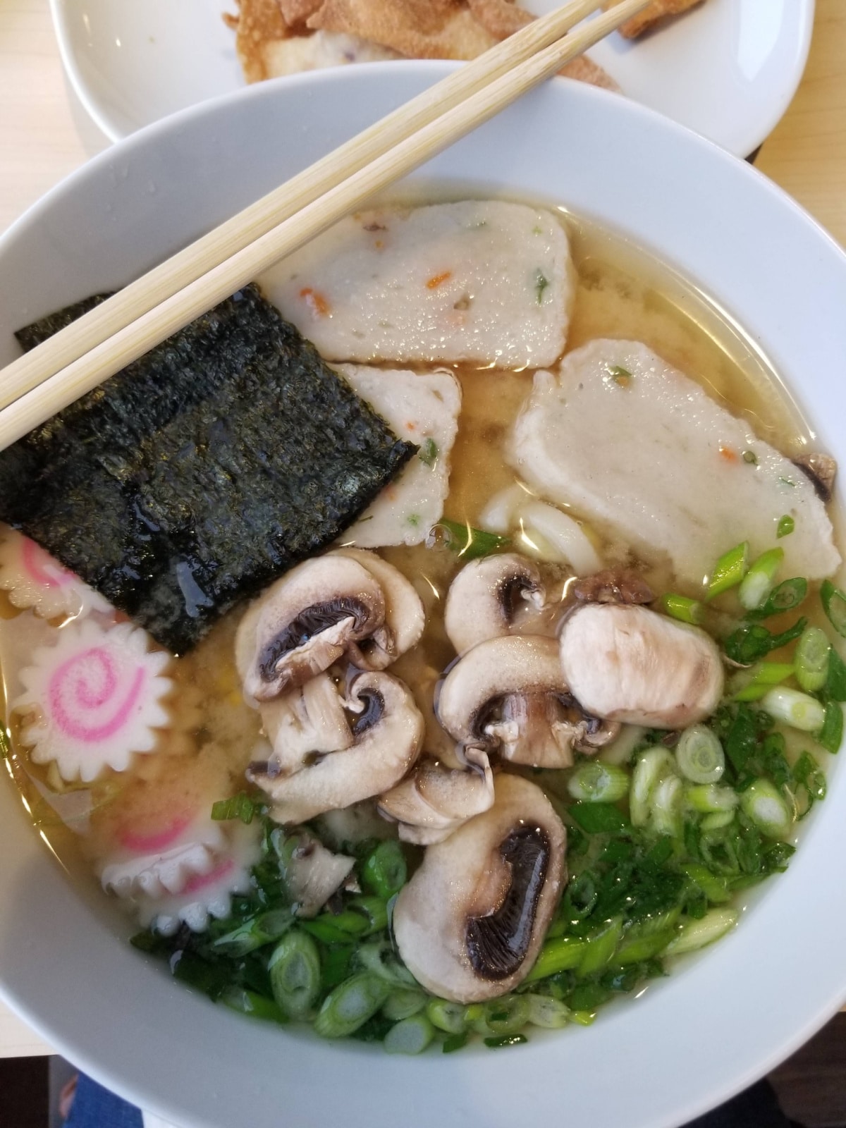 A bowl filled with broth, udon noodles, mushrooms, green onions, seaweed, and Narutomaki.
