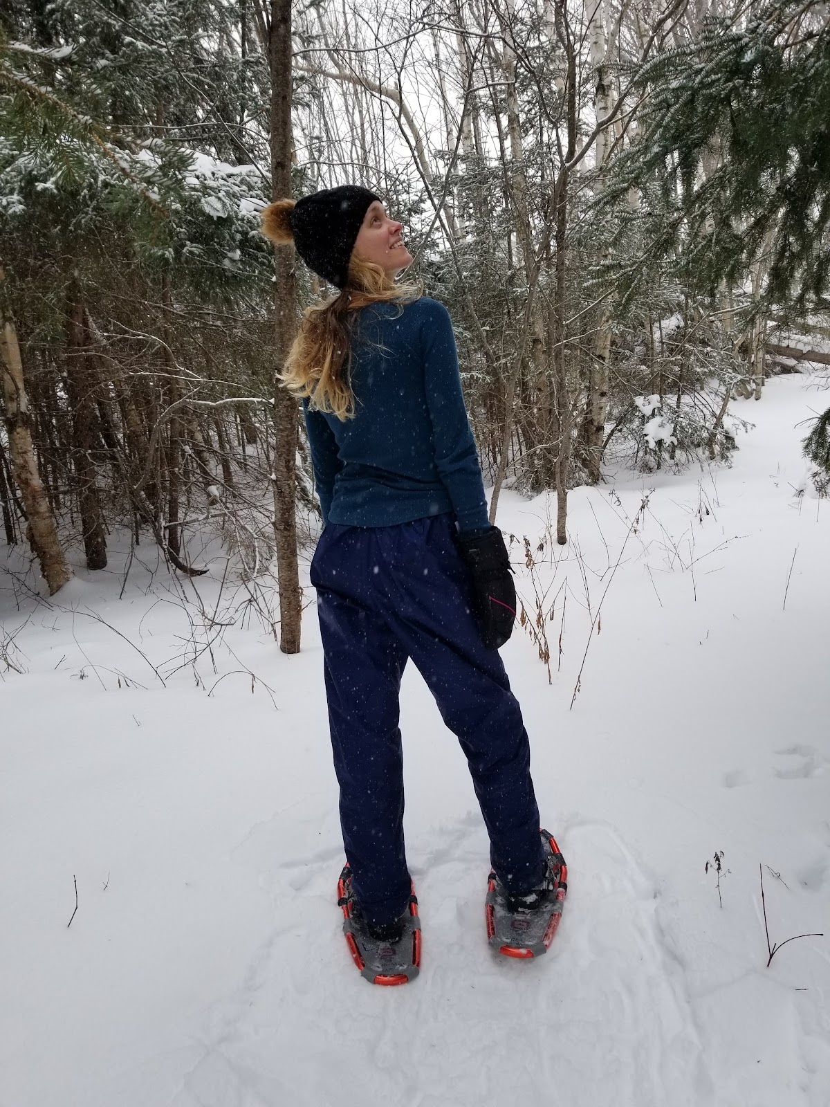 I am standing in the snow looking up at the sky. I am wearing a black hat, a green sweater, blue snow pants and snow shoes.