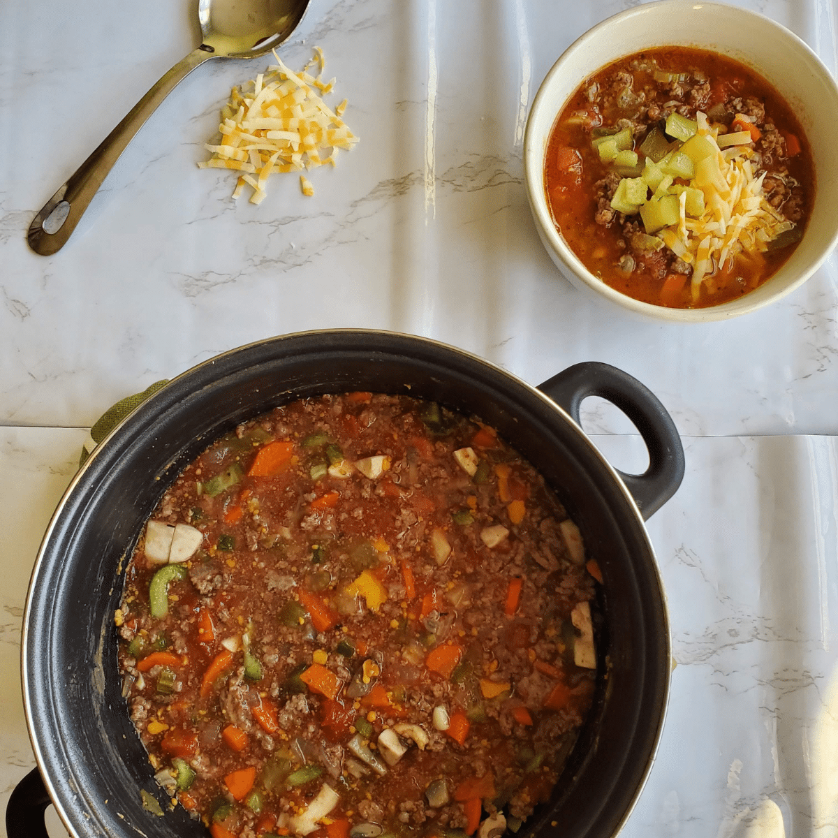 A pot of soup in the center of the photo with a spoon and shredded cheese in the left hand corner. In the right corner is a bowl of soup garnished with sliced pickles and shredded cheese.