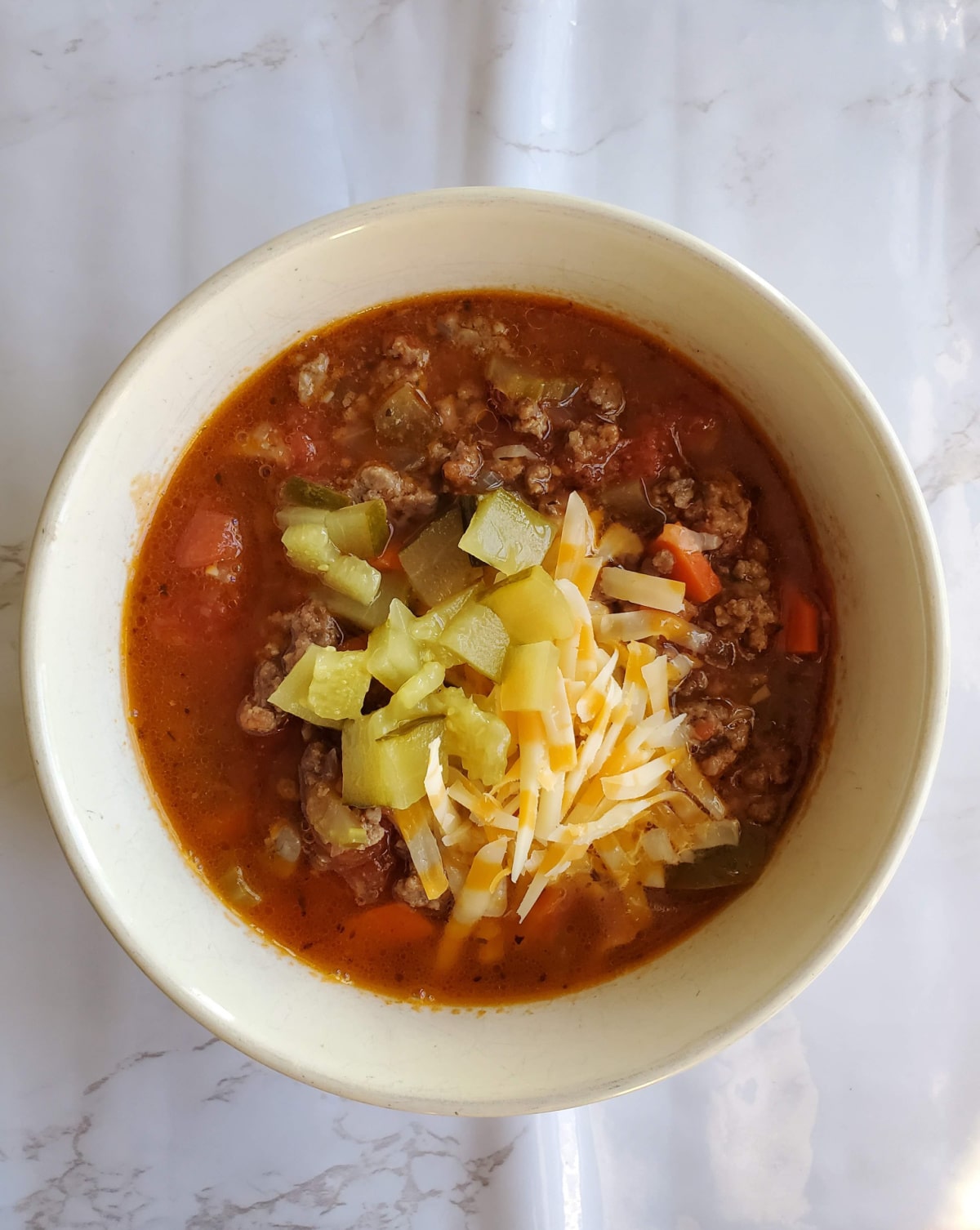 A bowl of hamburger vegetable soup garnished with chopped pickles and shredded cheese.