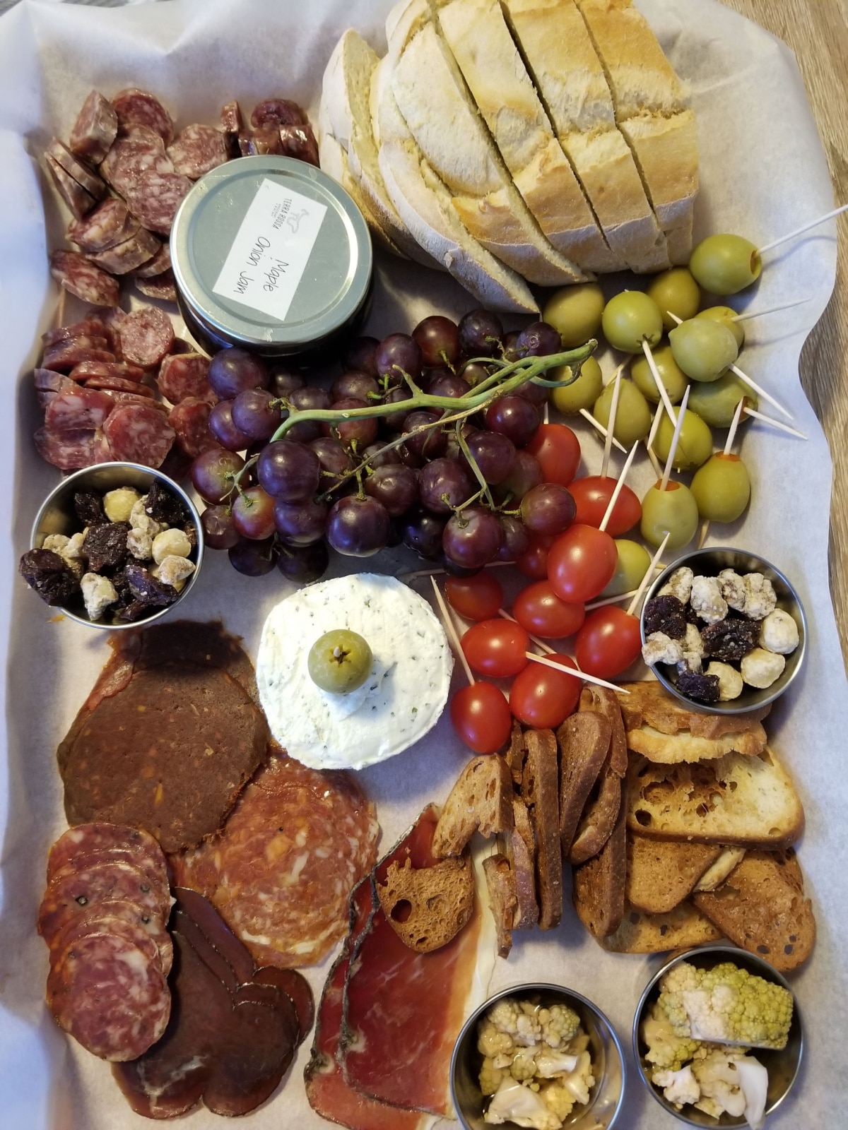 A charcuterie board full of green olives, grapes, cherry tomatoes, cheese, meats, sliced bread, little dishes of nuts and raisins and a little jar of maple onion jam.
