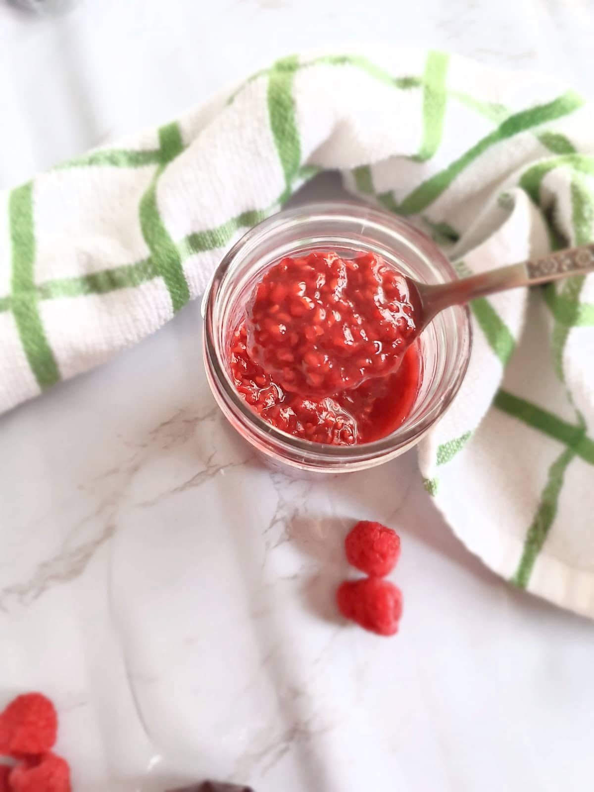 A mason jar full of raspberry compote. A spoon is in the jar lifting out some of the jam. A green and white linen is framing the jar and there are some raspberries around the jar. 