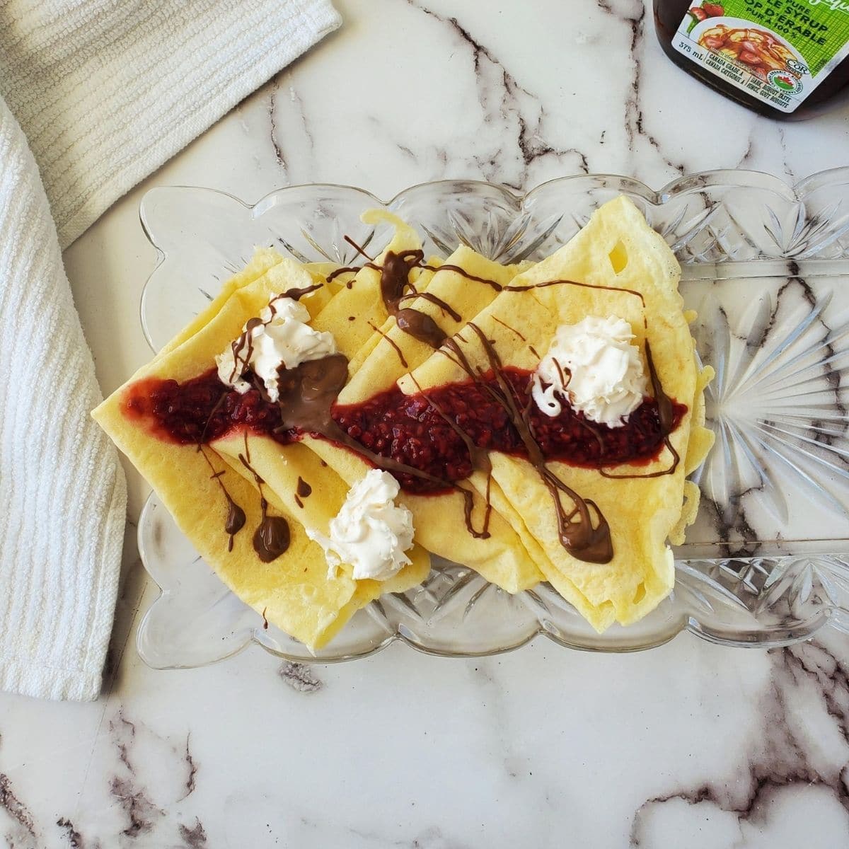 A glass serving dish with Belgian pancakes on it, topped with raspberry compote, melted chocolate and whipped cream. A bottle of syrup is in the right hand corner of the photo.