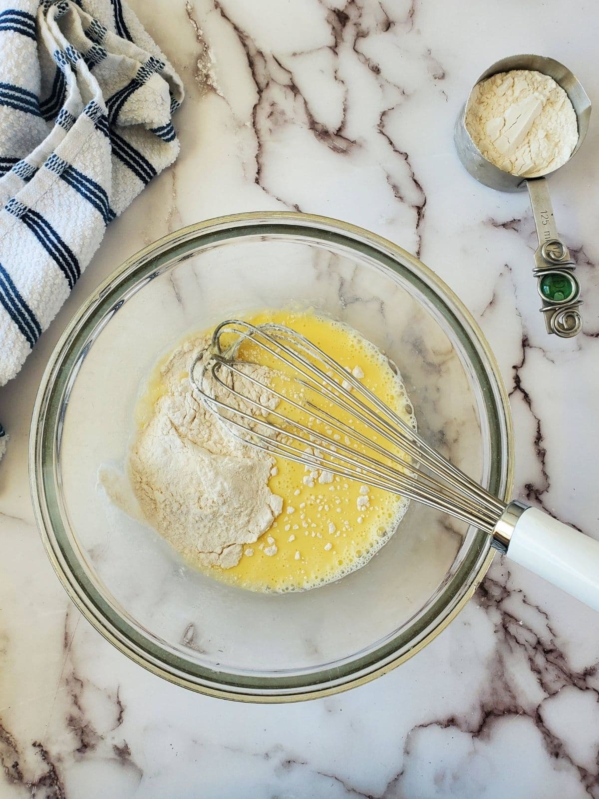 A glass mixing bowl with milk, egg and flour are in the center of the photo with a white and blue striped linen in the left hand corner. In the right hand corner there is a measuring cup with some white flour in it.