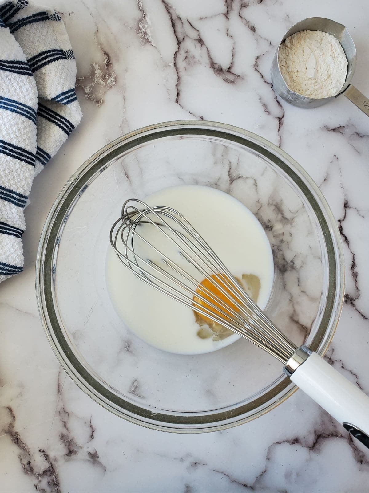 A glass mixing bowl with milk and an egg are in the center of the photo with a white and blue striped linen in the left hand corner. In the right hand corner there is a measuring cup with some white flour in it.