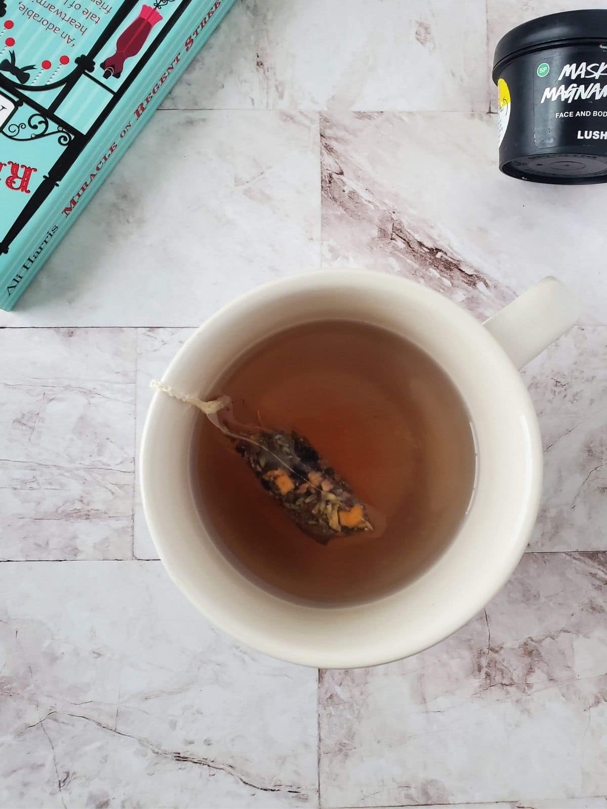 A mug of tea in the center of the photo and around it is a book and a face mask.