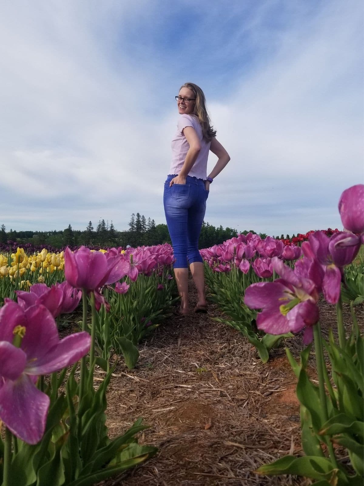 A blond girl is standing in a tulip field smiling at the camera. She is wearing a purple top and blue cropped jeans.