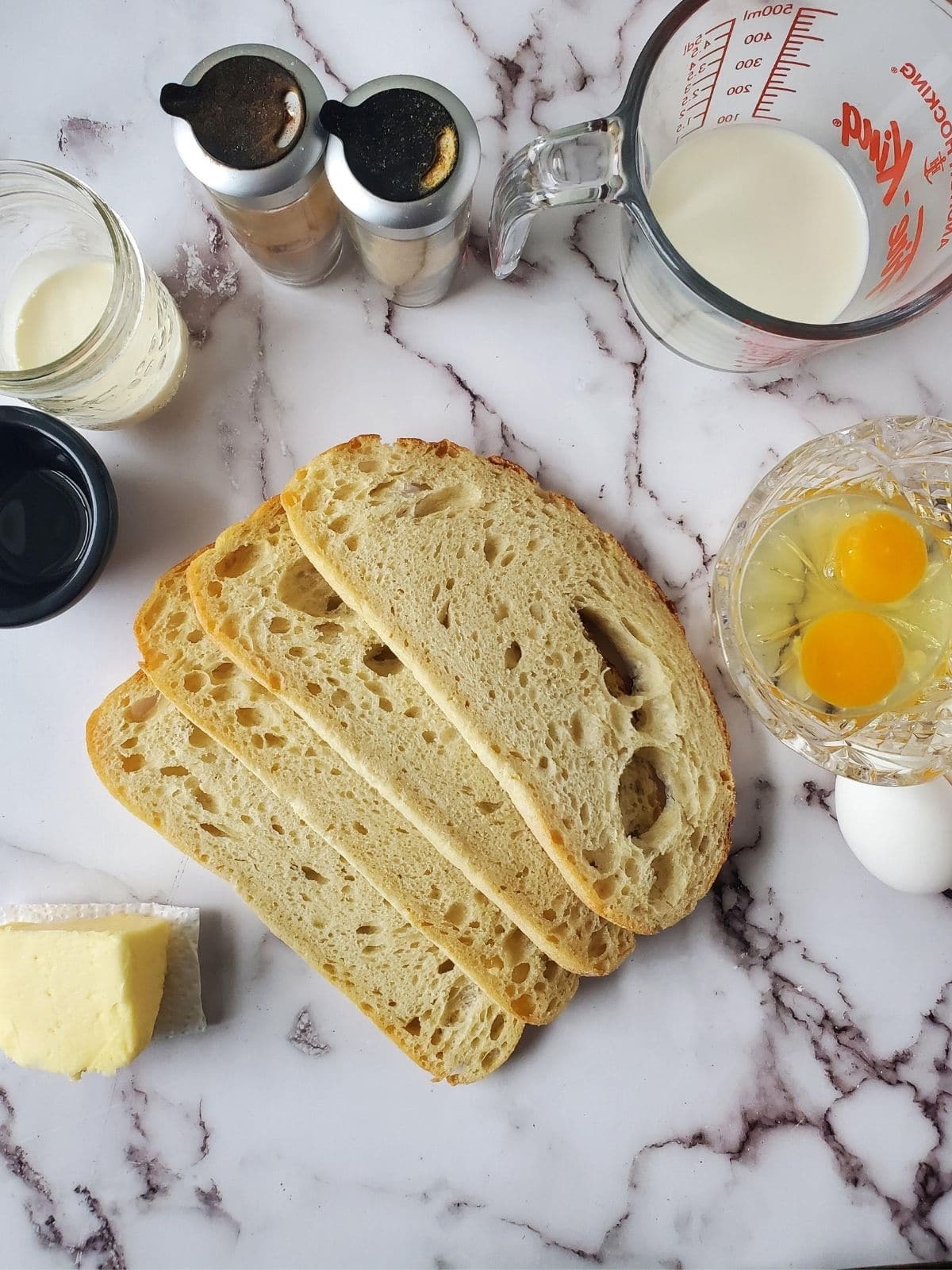 There are 4 slices of sourdough bread in the center of the photo. Around them are glasses dishes of cinnamon, nutmeg, milk, heavy cream, vanilla, and eggs. There is also a little stick of butter.