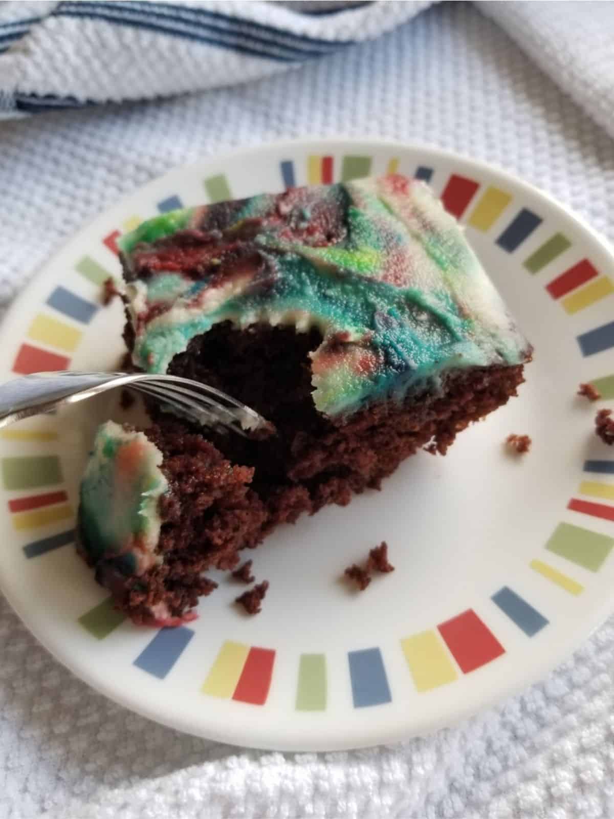 A slice of vegan chocolate cake with colorful icing. There is a fork taking a piece out of the cake.