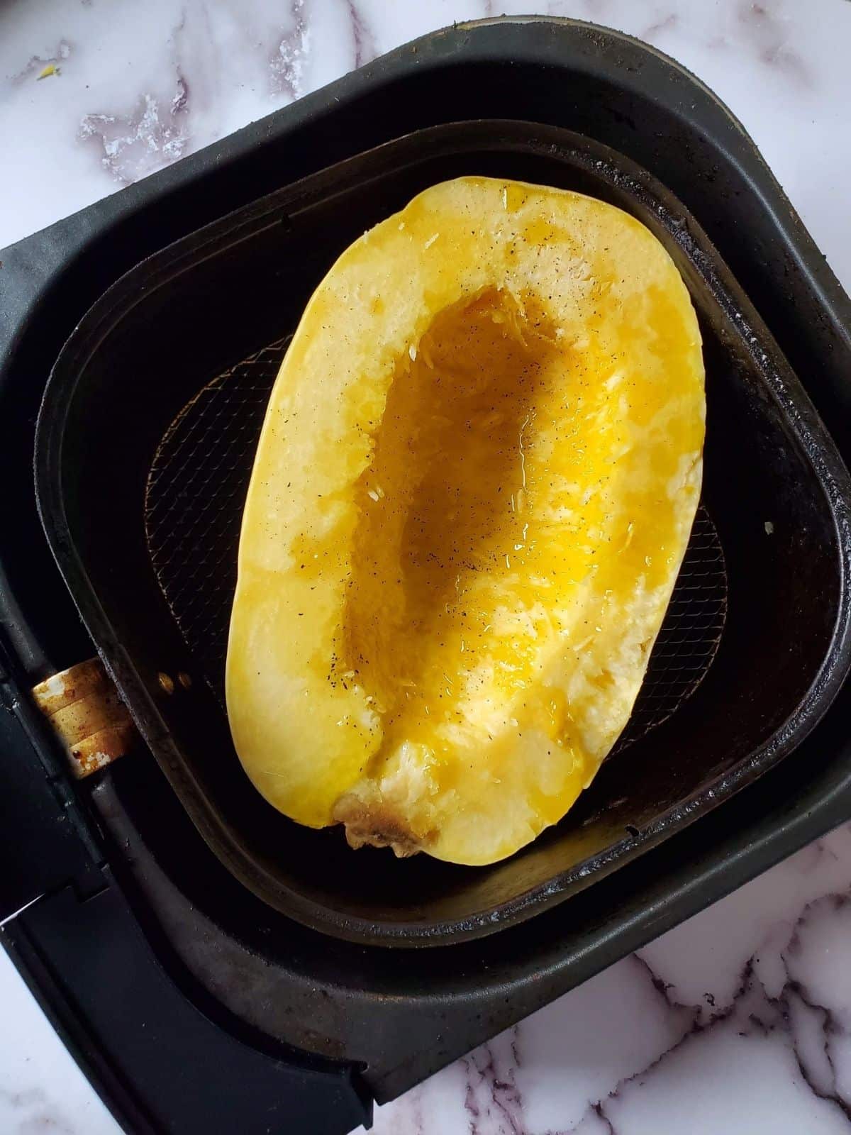 Half of a spaghetti squash sitting in an air fryer basket with olive oil and salt and pepper on it.