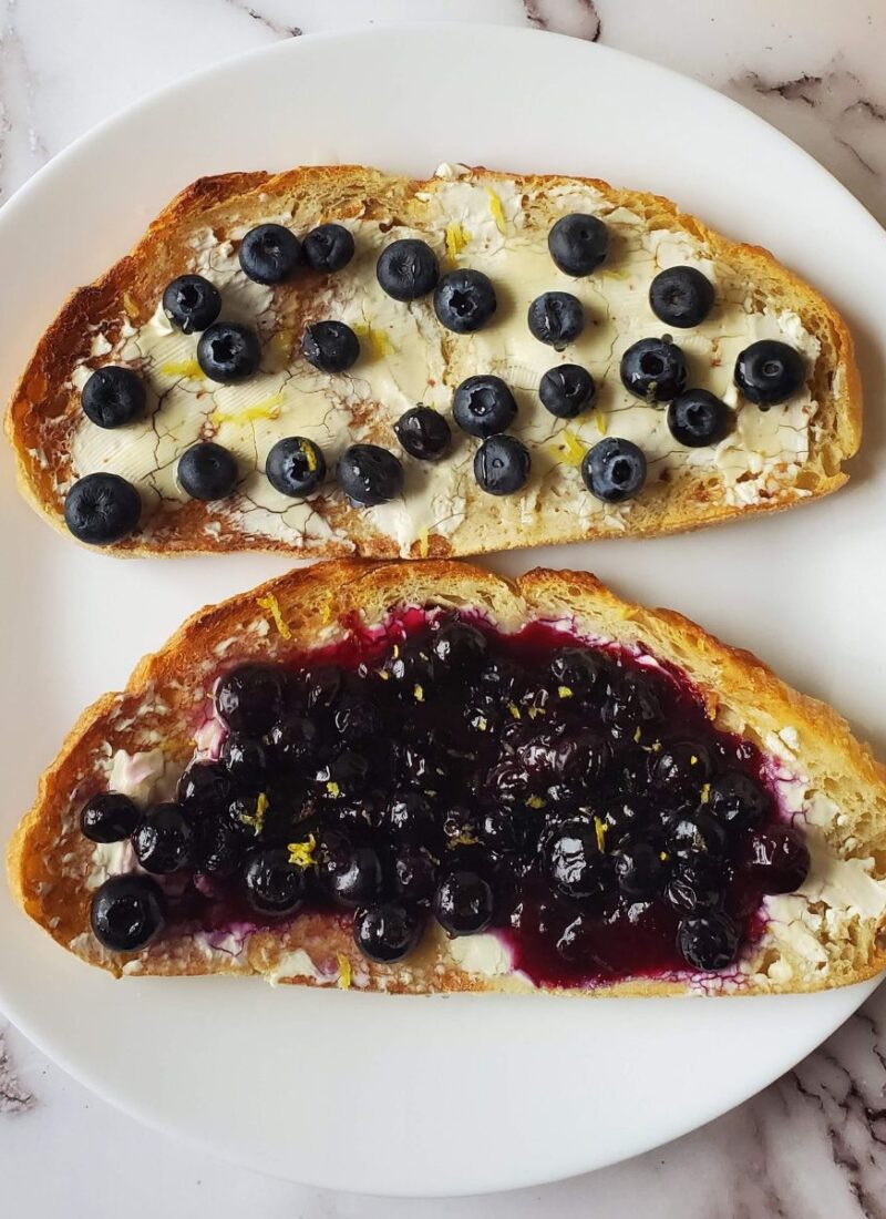 Two slices of blueberry toast. They both have cream cheese but one has whole blueberries on it the other one has homemade blueberry jam on it.