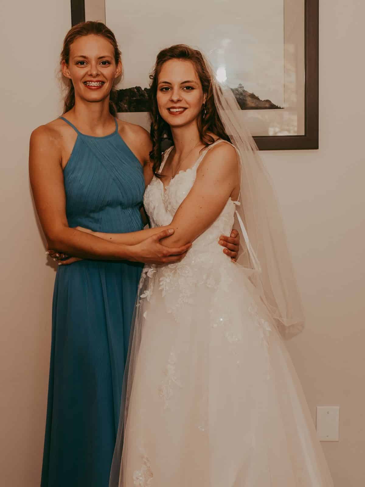 Two girls standing together. One is wearing a wedding dress the other a blue bridesmaid dress.