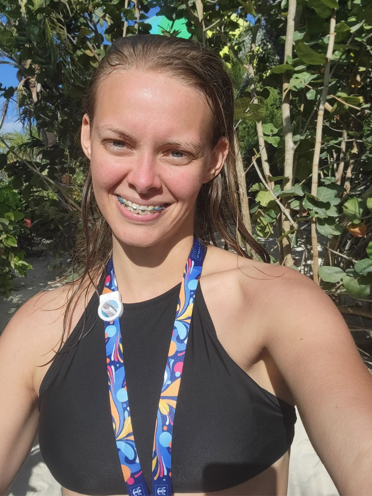 A blond girl is smiling at the camera wearing a black swim top and a lanyard around her neck. Her hair is damp and her face is red from the sun.