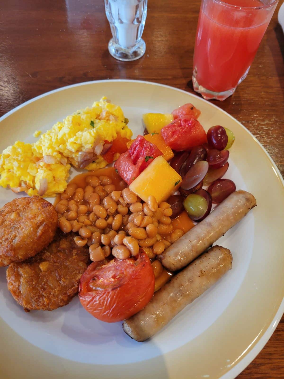 A plate of beans, sausages, scrambled eggs, hash browns, tomatoes, and some fruit. 