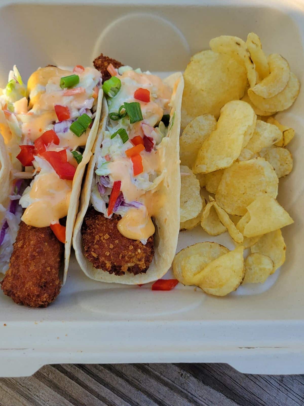 A takeout tray with two panko crusted mahi fish tacos with a side of potato chips. The tacos are topped with green onions and pepper and a creamy coleslaw and aioli dressing.