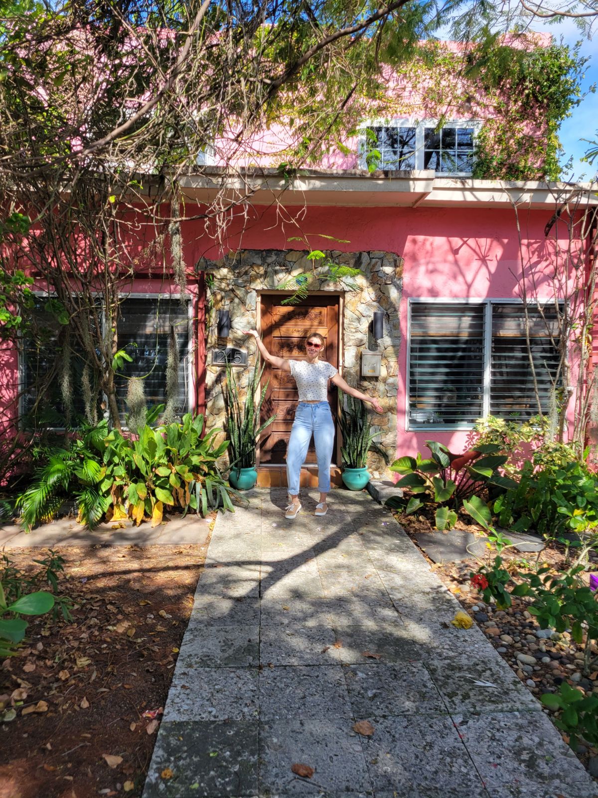 A blonde girl standing outside of an air bnb. The house is pink with a stone doorway and a wooden door. There is greenery growing around the pathway.