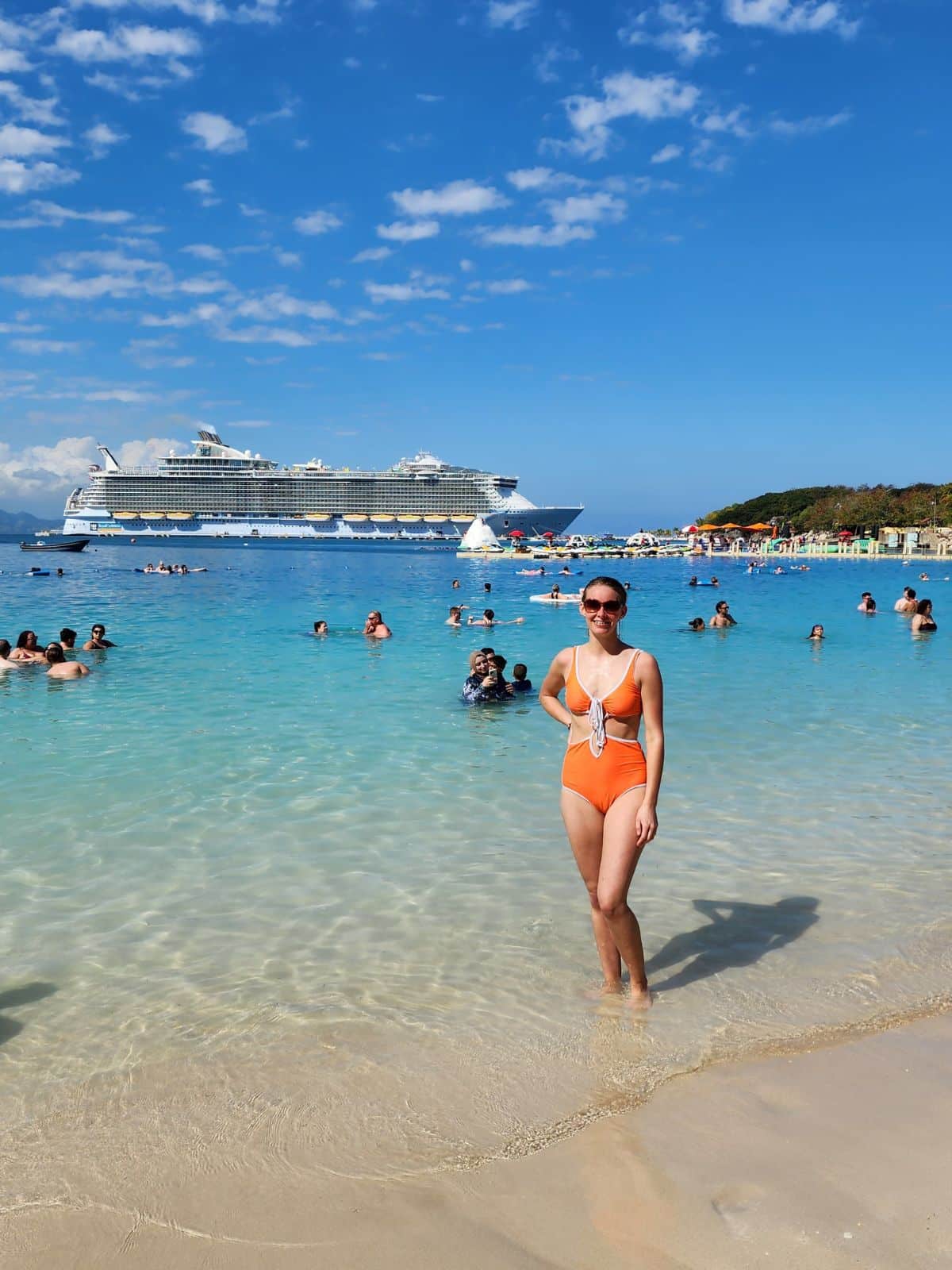 A girl is standing on the shore and there are people swimming in the ocean behind her. You can see the Oasis of the Seas cruise ship in the background.